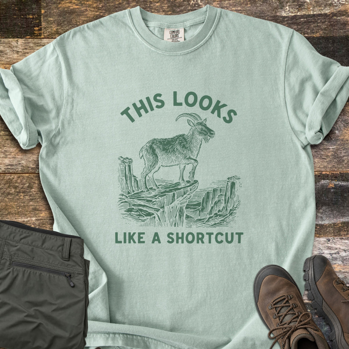 This Looks Like A Shortcut Comfort Colors T-shirt in Bay, laid flat. A dark green etching of a horned goat standing on a cliff ledge over a canyon, with the words This Looks arched above and Like A Shortcut below. Olive hiking pants and brown hiking boots lie beside it on dark wood.