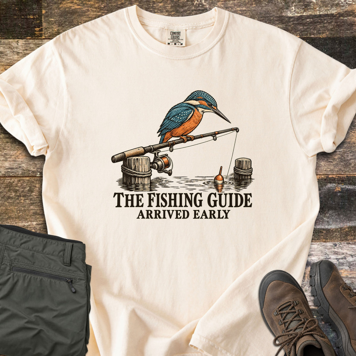 The Fishing Guide Arrived Early Comfort Colors T-shirt in Ivory, laid flat. A kingfisher perches on a fishing rod resting on rope-wrapped dock posts, its line and orange bobber in the water, above the words The Fishing Guide Arrived Early. Olive pants and brown hiking boots lie on weathered wood.