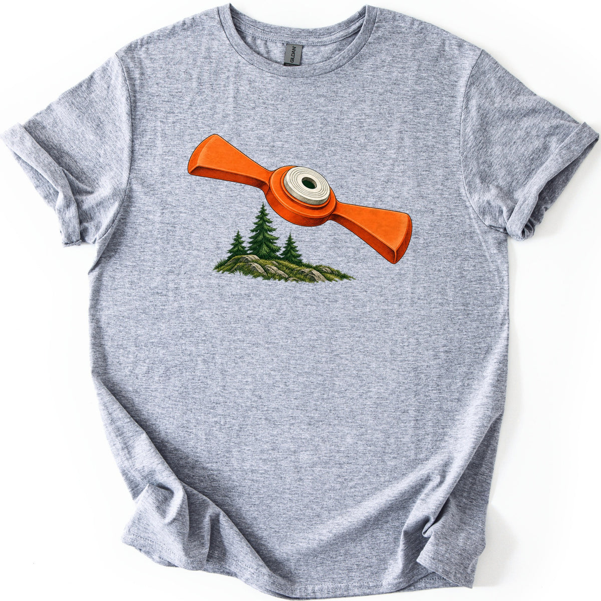 Winged Target Over the Pines Comfort Colors T-shirt in Sport Grey, laid flat. An orange clay target with flat, flared wings and a white center ring soars over three pines on a mossy rock outcrop, with no text. The heathered shirt lies on white with cuffed sleeves.