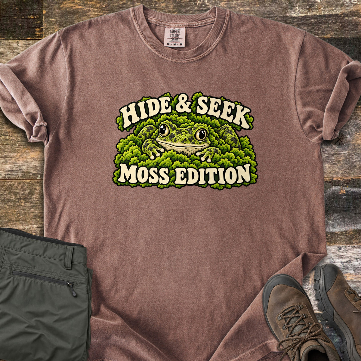 Hide & Seek Moss Edition Comfort Colors T-shirt in Espresso, laid flat. A wide-eyed frog peeks out from a mound of green moss between the words Hide & Seek and Moss Edition. Olive cargo pants and brown hiking boots lie beside it on weathered wood planks.