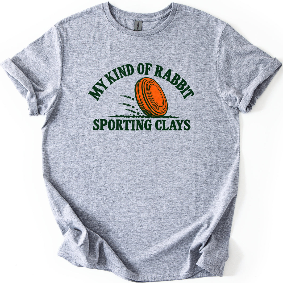 My Kind of Rabbit Comfort Colors T-shirt in Sport Grey, laid flat. An orange clay target rolls through grass with motion lines, framed by the arched words My Kind of Rabbit above and Sporting Clays below in dark green. The shirt has rolled sleeves on a white background.