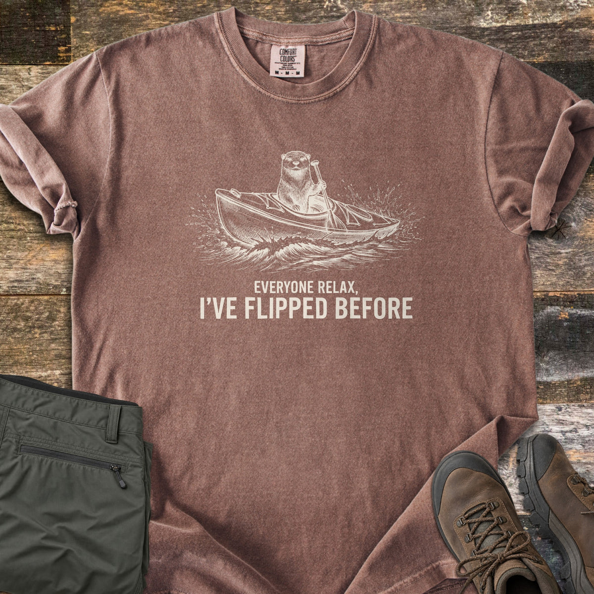 Everyone Relax I've Flipped Before Comfort Colors T-shirt in Espresso, laid flat. A cream sketch of an otter paddling a kayak through splashing waves above the words Everyone Relax, I've Flipped Before. Sleeves are rolled, with olive hiking pants and brown boots on rustic wood planks.