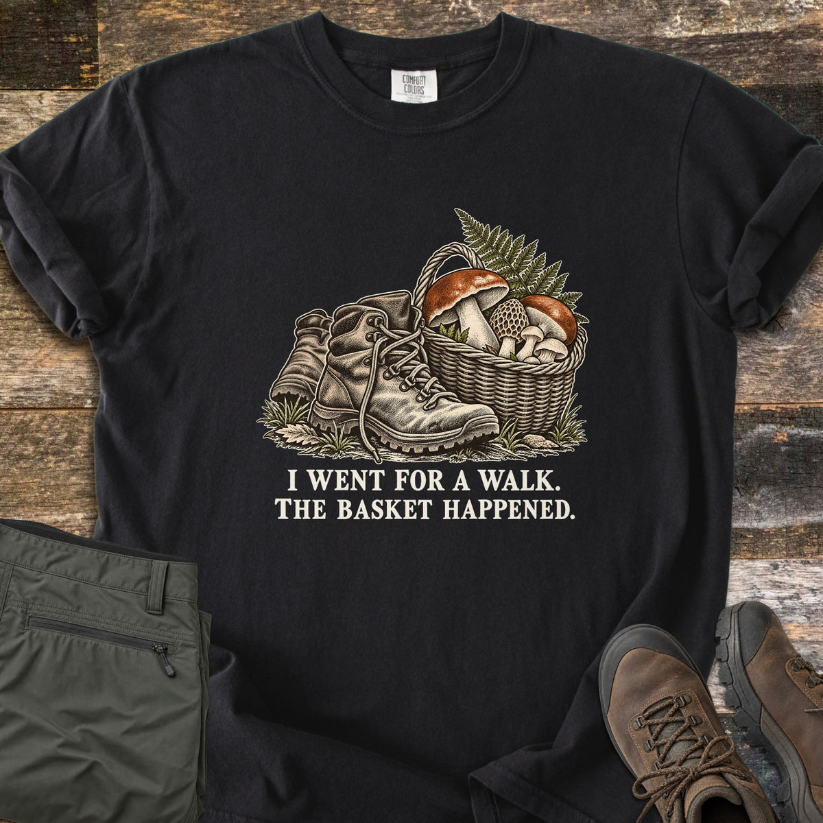 I Went for a Walk. The Basket Happened Comfort Colors T-shirt in Black, laid flat. A vintage-style illustration of worn hiking boots beside a wicker basket of mushrooms and fern, above the words I Went for a Walk. The Basket Happened in cream. On rustic wood with olive cargo pants and brown hiking boots.