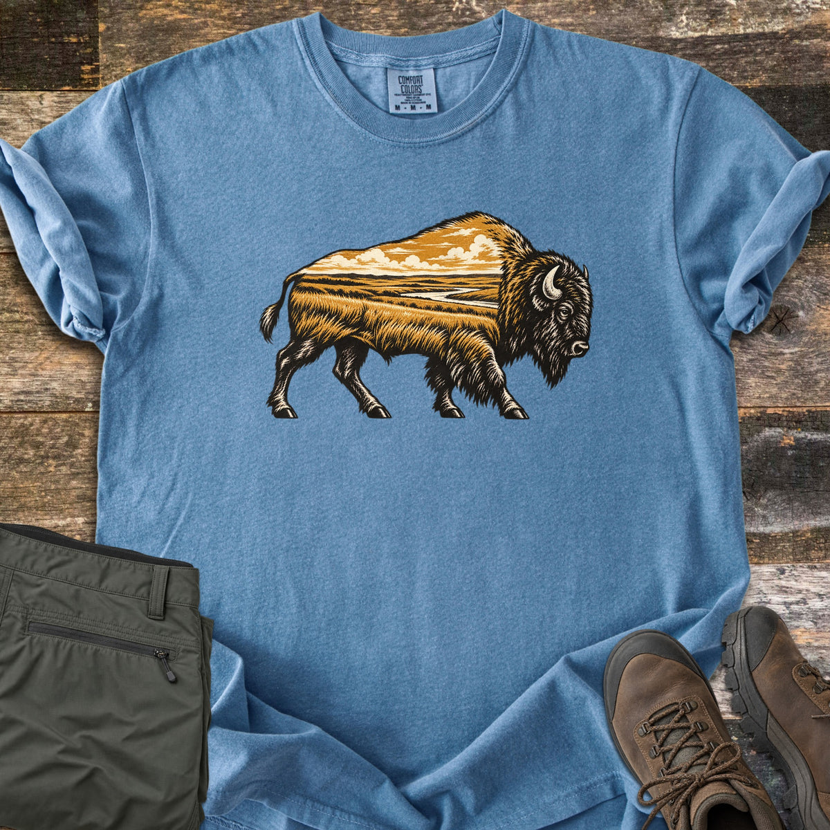 Prairie Bison Comfort Colors T-shirt in Blue Jean, laid flat. A walking bison in black woodcut-style linework, its body filled with a golden prairie scene of grassland, a winding river and clouds. Folded olive cargo pants and brown hiking boots sit on weathered wood.