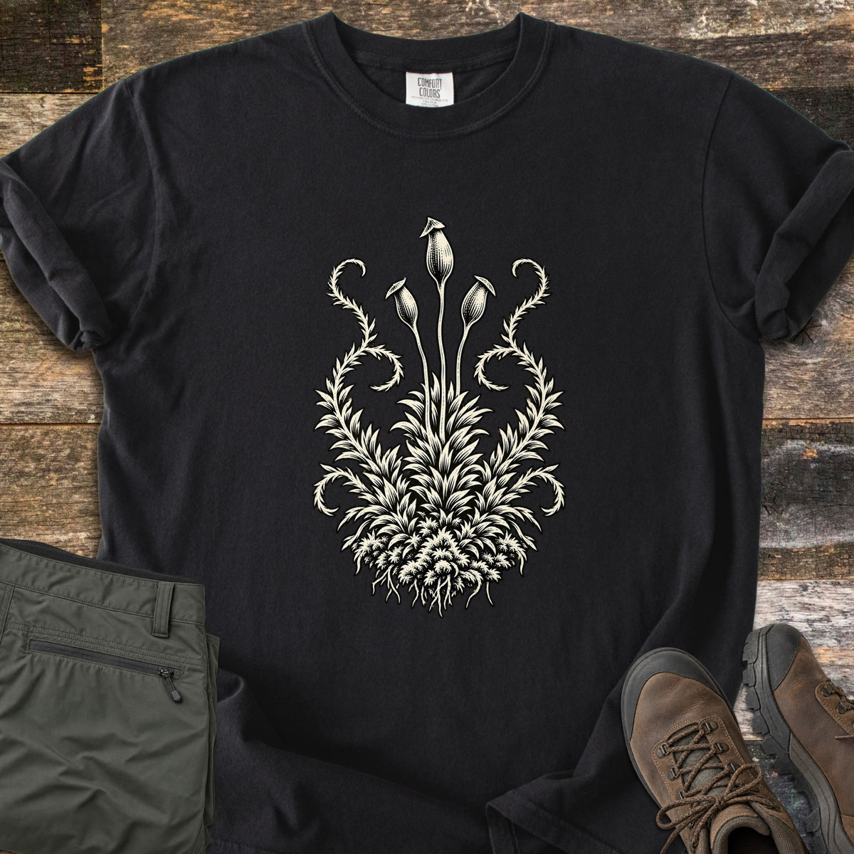 Moss Woodcut Comfort Colors T-shirt in Black, laid flat. A cream woodcut-style print of a moss clump with three tall spore capsules on stems and curling fronds on each side. Olive cargo pants and brown hiking boots sit beside it on weathered wood planks.