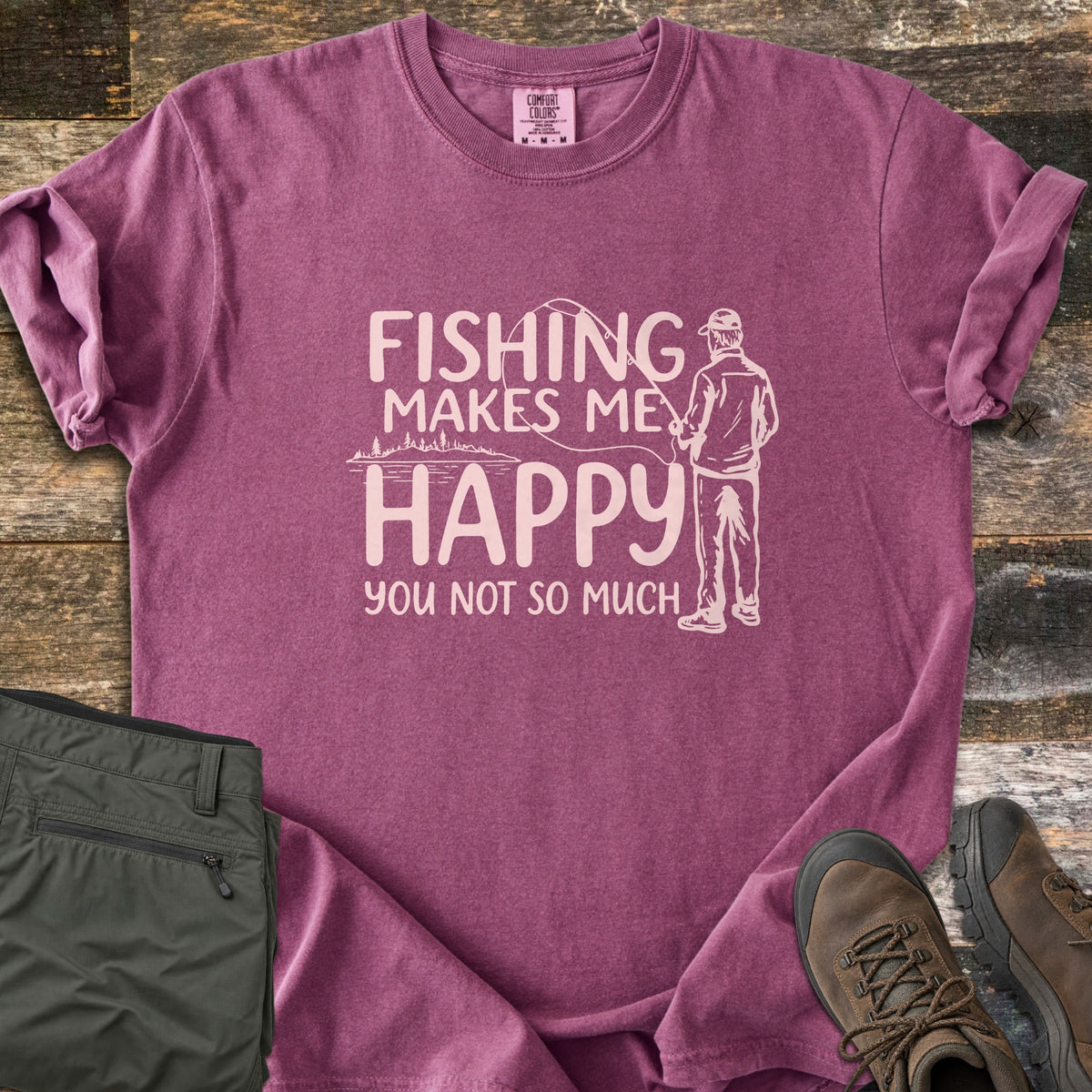 Fishing Makes Me Happy Comfort Colors T-shirt in Berry, laid flat. Light pink print of a man in a cap casting a fishing rod by a tree-lined lake, with the words Fishing Makes Me Happy You Not So Much. Laid on weathered wood with olive pants and brown hiking boots.