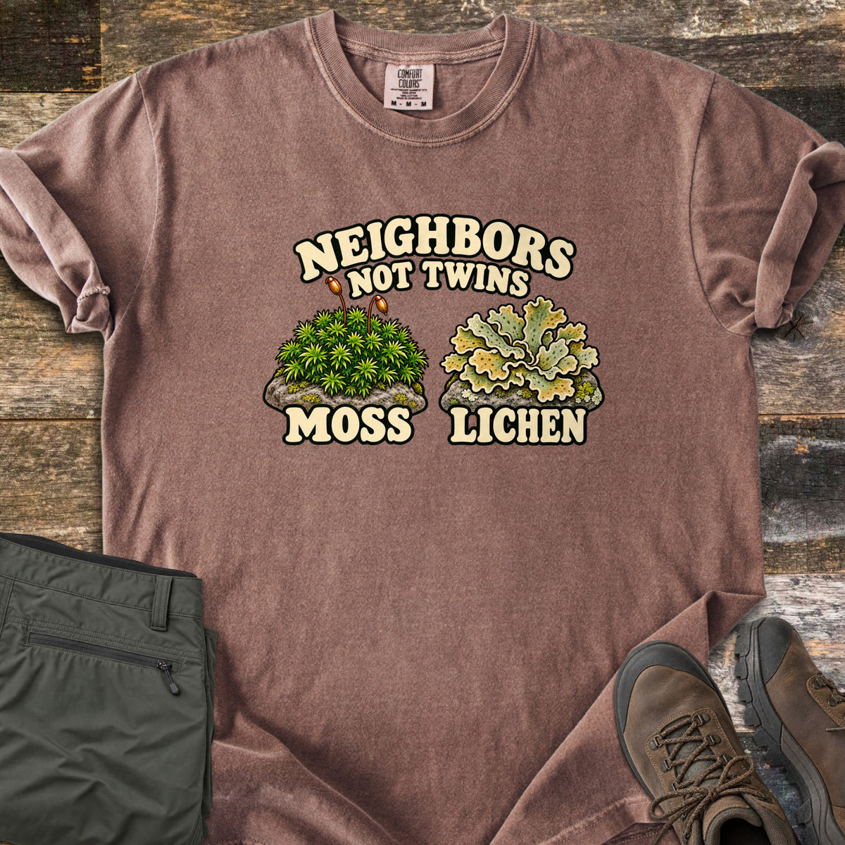 Neighbors Not Twins Comfort Colors T-shirt in Espresso, laid flat. Arched text Neighbors Not Twins over two illustrated rock clumps: green moss with brown stalks labeled Moss, and pale green leafy lichen labeled Lichen. Olive hiking pants and brown boots sit beside it on weathered wood.