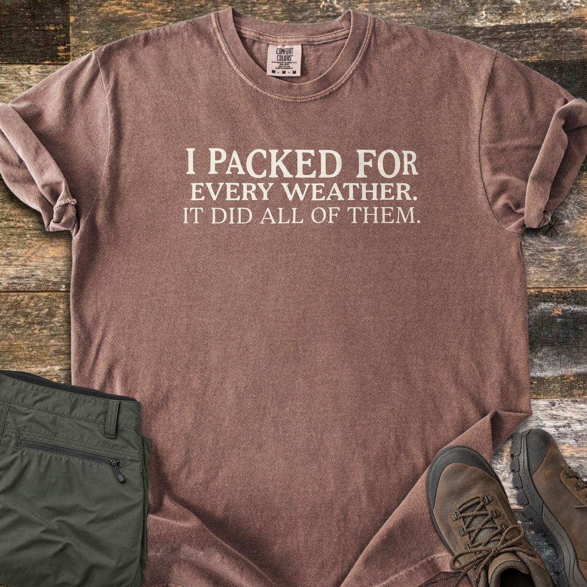 Packed For Every Weather Comfort Colors T-shirt in Espresso, laid flat. Cream serif text on the chest reads I PACKED FOR EVERY WEATHER. IT DID ALL OF THEM. The shirt lies on weathered wood planks with folded olive hiking pants and brown hiking boots.