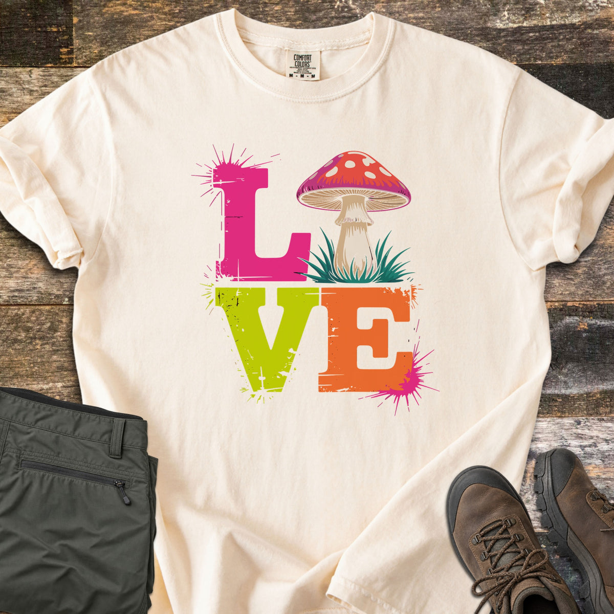 Mushroom Love Comfort Colors T-shirt in Ivory, laid flat. The word LOVE in distressed pink, lime green and orange letters with paint splatters, a red-capped mushroom on grass as the O. Shown on dark weathered wood with olive pants and brown hiking boots.