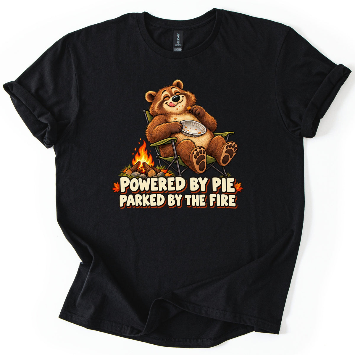 Powered by Pie Parked by the Fire Comfort Colors T-shirt in Black, laid flat. A smiling cartoon bear licks its lips in a green camp chair, holding an empty pie tin beside a campfire, above the words Powered by Pie Parked by the Fire. The shirt has rolled sleeves on a white background.