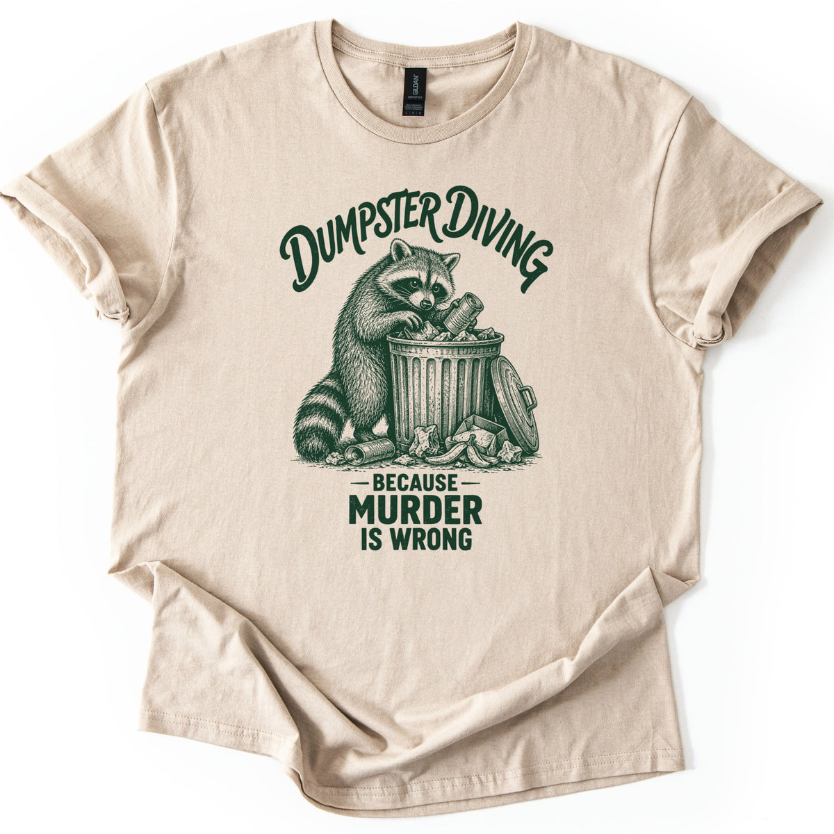 Dumpster Diving Murder Raccoon Comfort Colors T-shirt in Sand, laid flat. Dark green etching of a raccoon pulling a can from an overflowing trash can under arched script reading Dumpster Diving, with Because Murder Is Wrong below. Sleeves rolled on a white background.