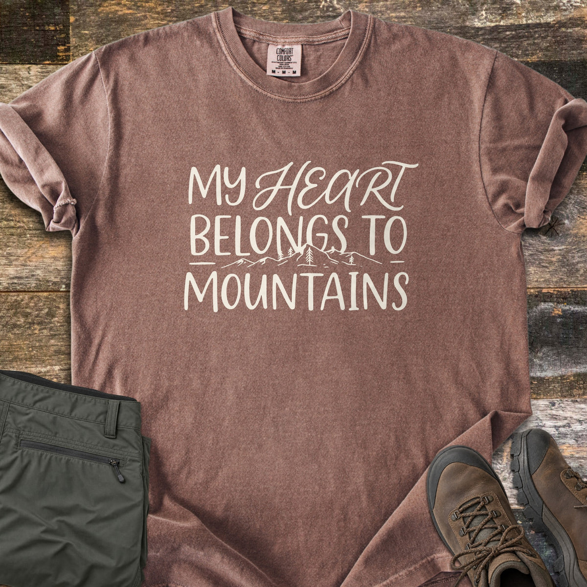 My Heart Belongs to Mountains Comfort Colors T-shirt in Espresso, laid flat. Cream hand lettering reads My Heart Belongs to Mountains, with Heart in script and a line drawing of peaks and pine trees. The sleeves are rolled, with olive pants and brown hiking boots on dark wood.