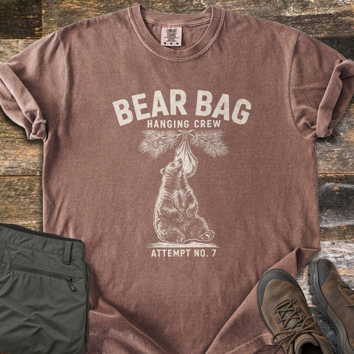 Bear Bag Hanging Crew Comfort Colors T-shirt in Espresso, laid flat. A sketched bear sits under a pine branch sniffing a hanging food bag, with the words Bear Bag Hanging Crew above and Attempt No. 7 below. Olive hiking pants and brown hiking boots lie beside it on weathered wood.