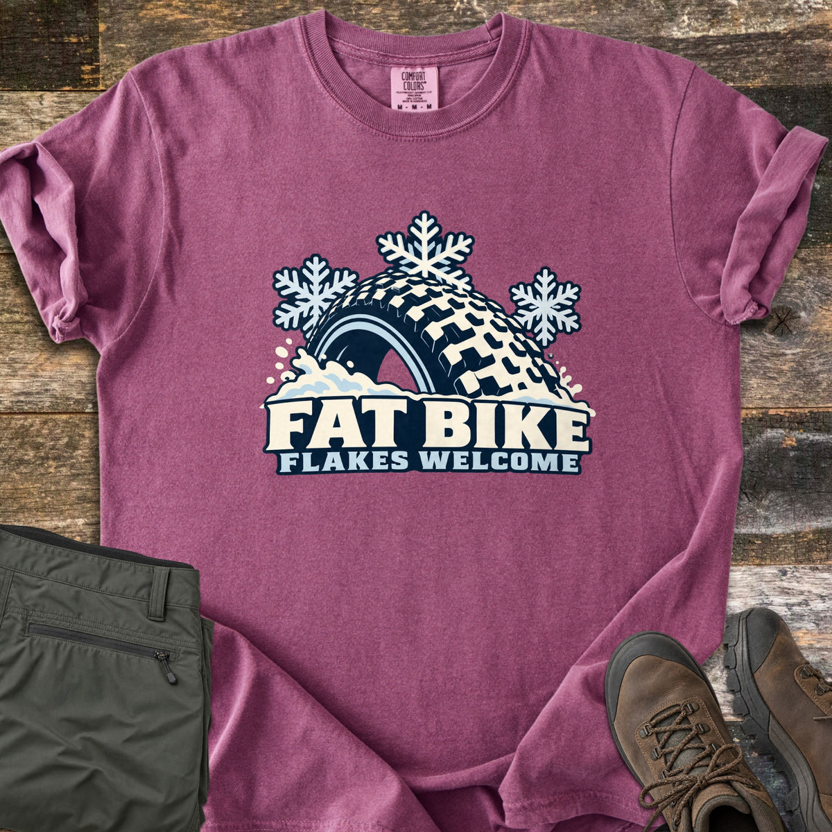 Fat Bike Flakes Welcome Comfort Colors T-shirt in Berry, laid flat. A fat bike tire half buried in snow with three snowflakes above it, over the words FAT BIKE FLAKES WELCOME. Sleeves are rolled, on a wood floor with folded olive pants and brown hiking boots.
