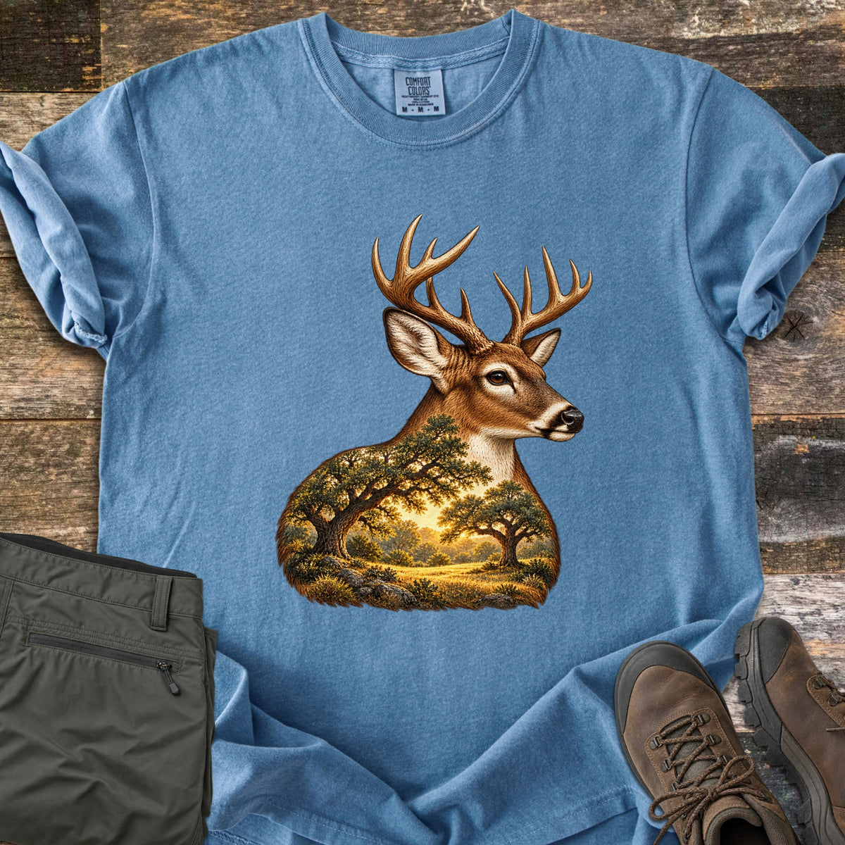 Whitetail Oak Grove Comfort Colors T-shirt in Blue Jean, laid flat. A whitetail buck with branching antlers, its neck and chest filled with a sunlit grove of oak trees and grassy meadow. The shirt lies on weathered wood planks beside folded olive pants and brown hiking boots.