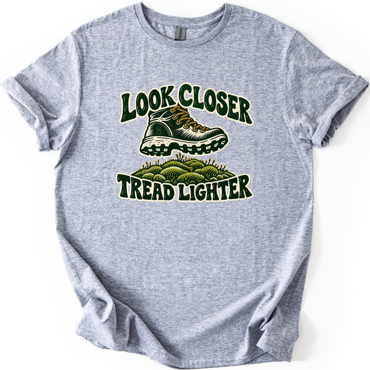 Look Closer Tread Lighter Comfort Colors T-shirt in Sport Grey, laid flat. A hiking boot with gold laces steps over a patch of green moss, with Look Closer arched above and Tread Lighter below in curvy dark green letters. The shirt is spread on a plain white surface with cuffed sleeves.