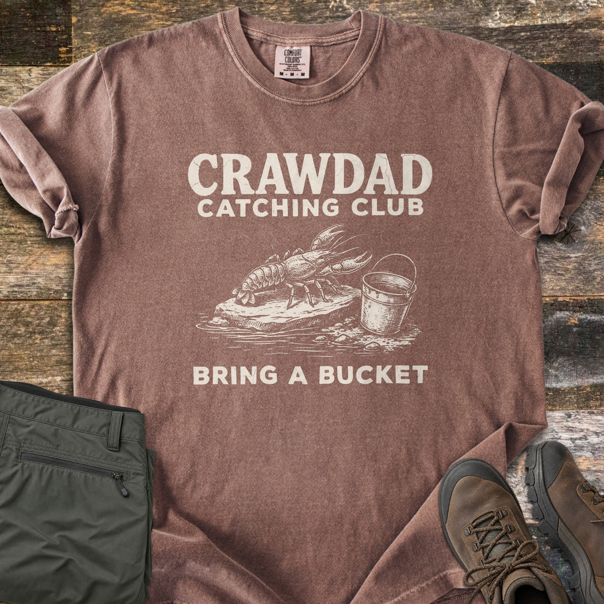 Crawdad Catching Club Comfort Colors T-shirt in Espresso, laid flat. Cream print reads CRAWDAD CATCHING CLUB above a sketch of a crawdad on a rock beside a bucket, with BRING A BUCKET below. Folded olive pants and brown hiking boots lie beside it on weathered wood.