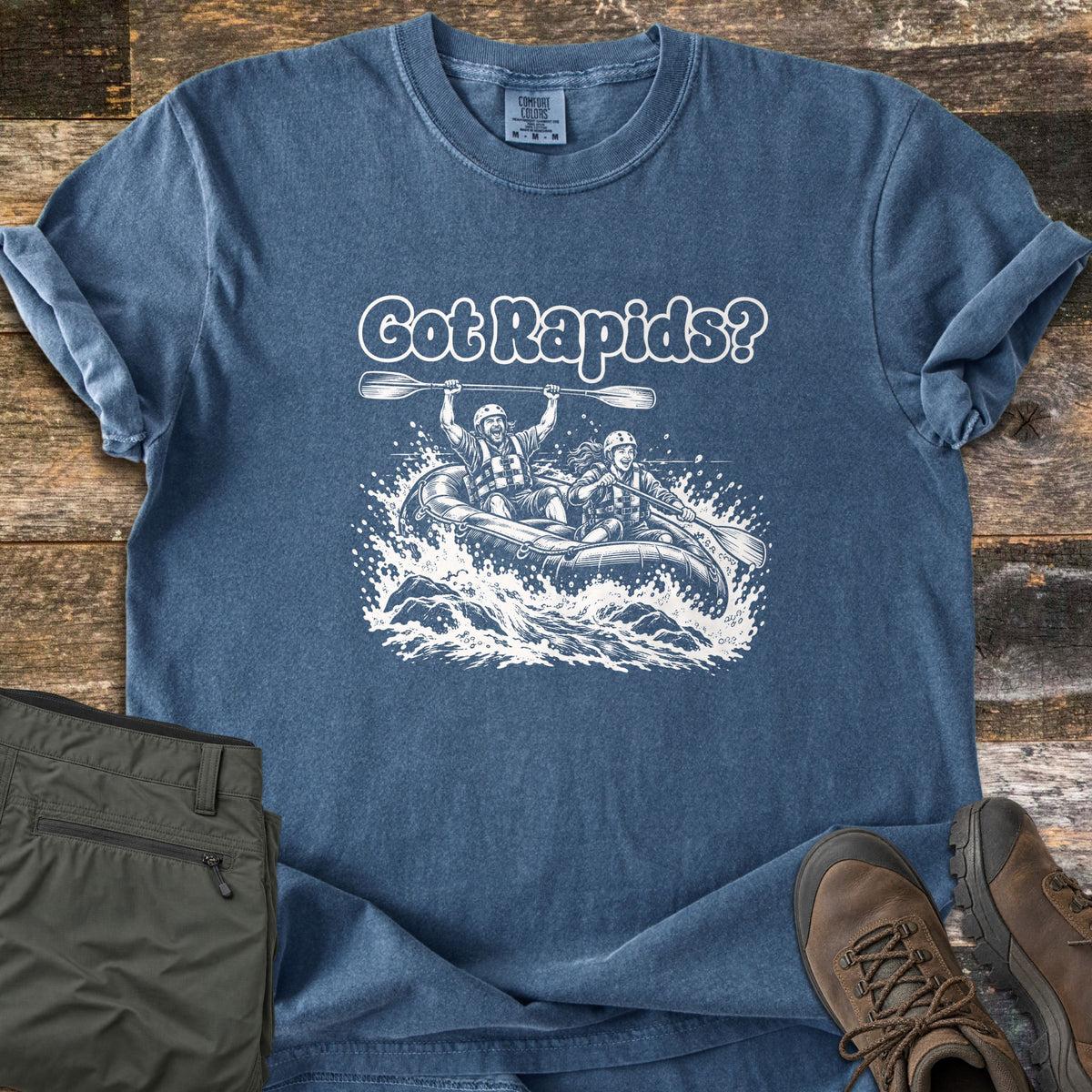 Got Rapids Comfort Colors T-shirt in Denim, laid flat. Printed in white: the words Got Rapids? above two smiling rafters in helmets and life vests paddling an inflatable raft through splashing whitewater. Olive pants and brown hiking boots sit on a wood floor.