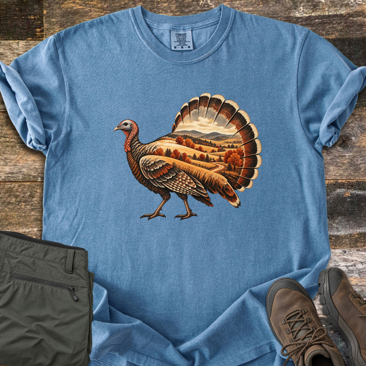 Turkey Harvest Hills Comfort Colors T-shirt in Blue Jean, laid flat. A walking wild turkey whose body and fanned tail hold an autumn landscape of rolling golden hills, orange trees, distant mountains and a dirt road. Laid on rustic wood beside olive pants and brown hiking boots.