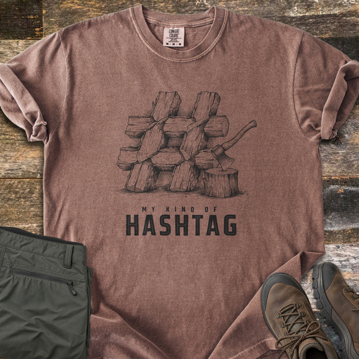 My Kind Of Hashtag Comfort Colors T-shirt in Espresso, laid flat. A sketched stack of split firewood logs arranged in a hashtag shape, with an axe in a stump, above the words My Kind Of Hashtag. Olive hiking pants and brown hiking boots lie beside it on weathered wood planks.