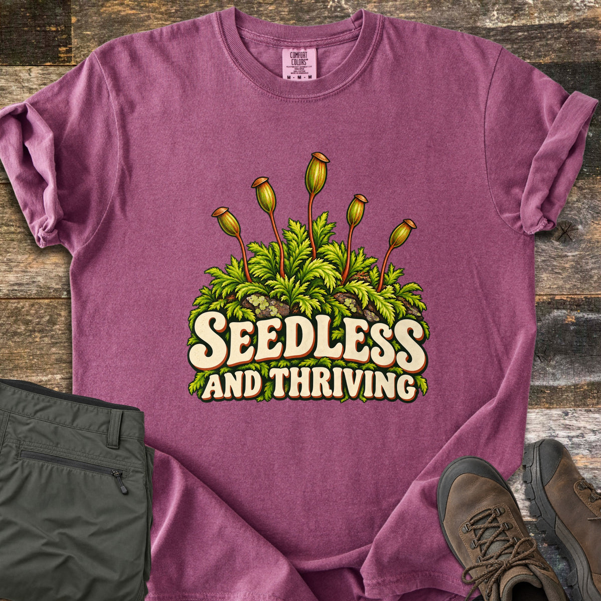 Seedless and Thriving Comfort Colors T-shirt in Berry, laid flat. Green feathery moss on lichen-covered rocks sends up tall stalks with spore capsules above the words Seedless and Thriving in cream retro letters. It lies on weathered wood beside olive pants and brown hiking boots.