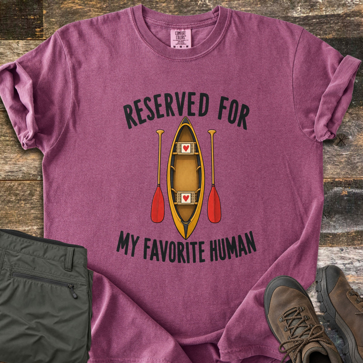 Reserved For My Favorite Human Comfort Colors T-shirt in Berry, laid flat. A yellow canoe seen from above with a red heart on each seat sits between two red-bladed paddles, under the words Reserved For and above My Favorite Human. Olive pants and brown hiking boots lie beside it on wood planks.