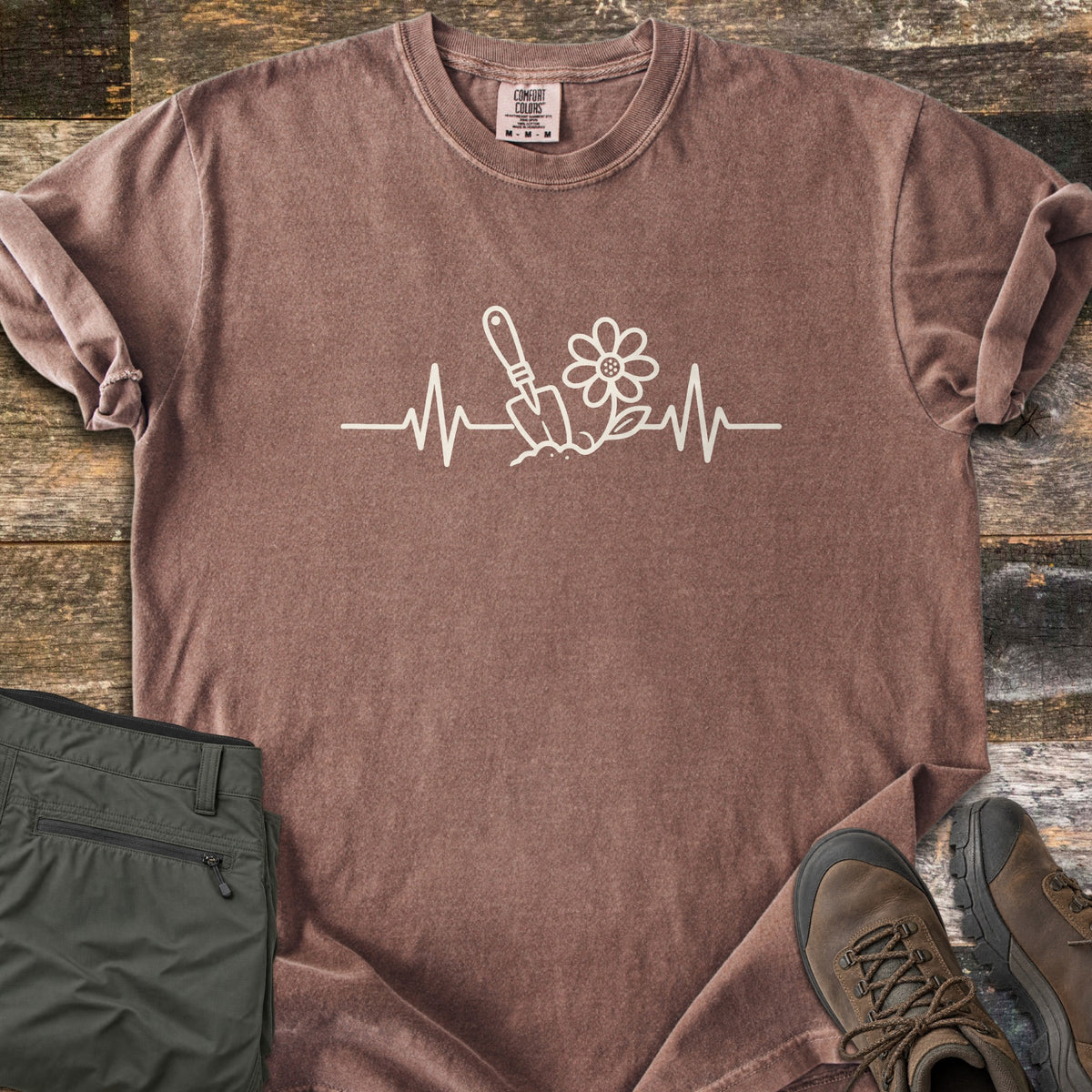 Gardening Heartbeat Comfort Colors T-shirt in Espresso, laid flat. A cream line drawing of a heartbeat line with a garden trowel in soil and a daisy at its center, with no words. Folded olive pants and brown hiking boots sit beside the shirt on weathered wood boards.