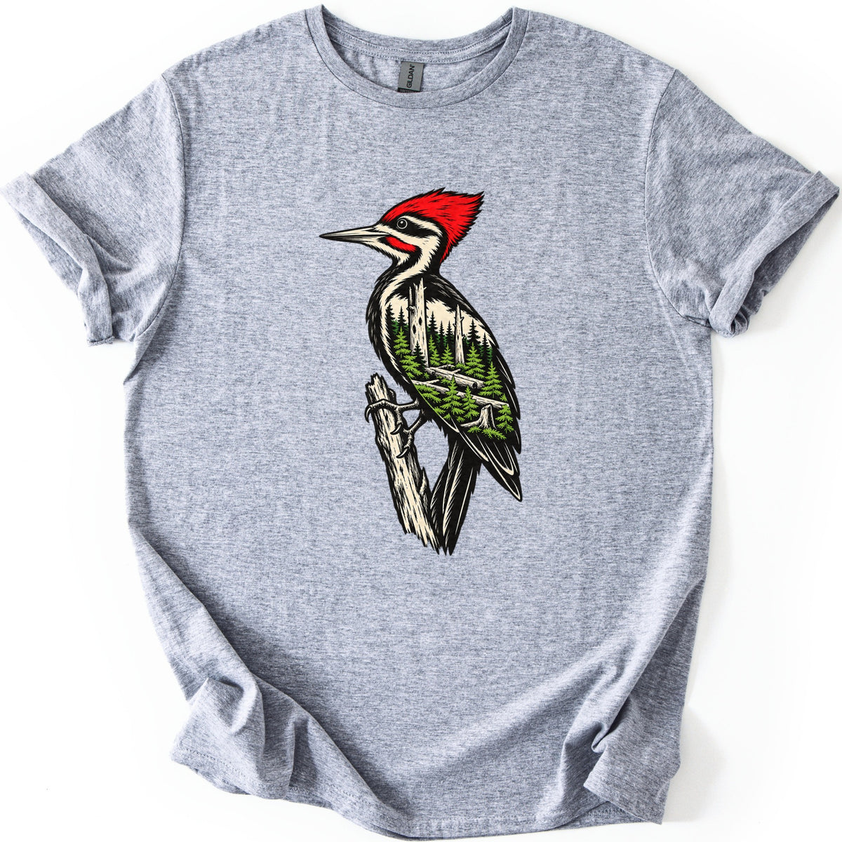 Woodpecker Forest Comfort Colors T-shirt in Sport Grey, laid flat. A red-crested woodpecker grips a weathered branch, its black and cream body filled with a pine forest and fallen logs. No words are printed. The sleeves are rolled and the shirt lies on a white background.