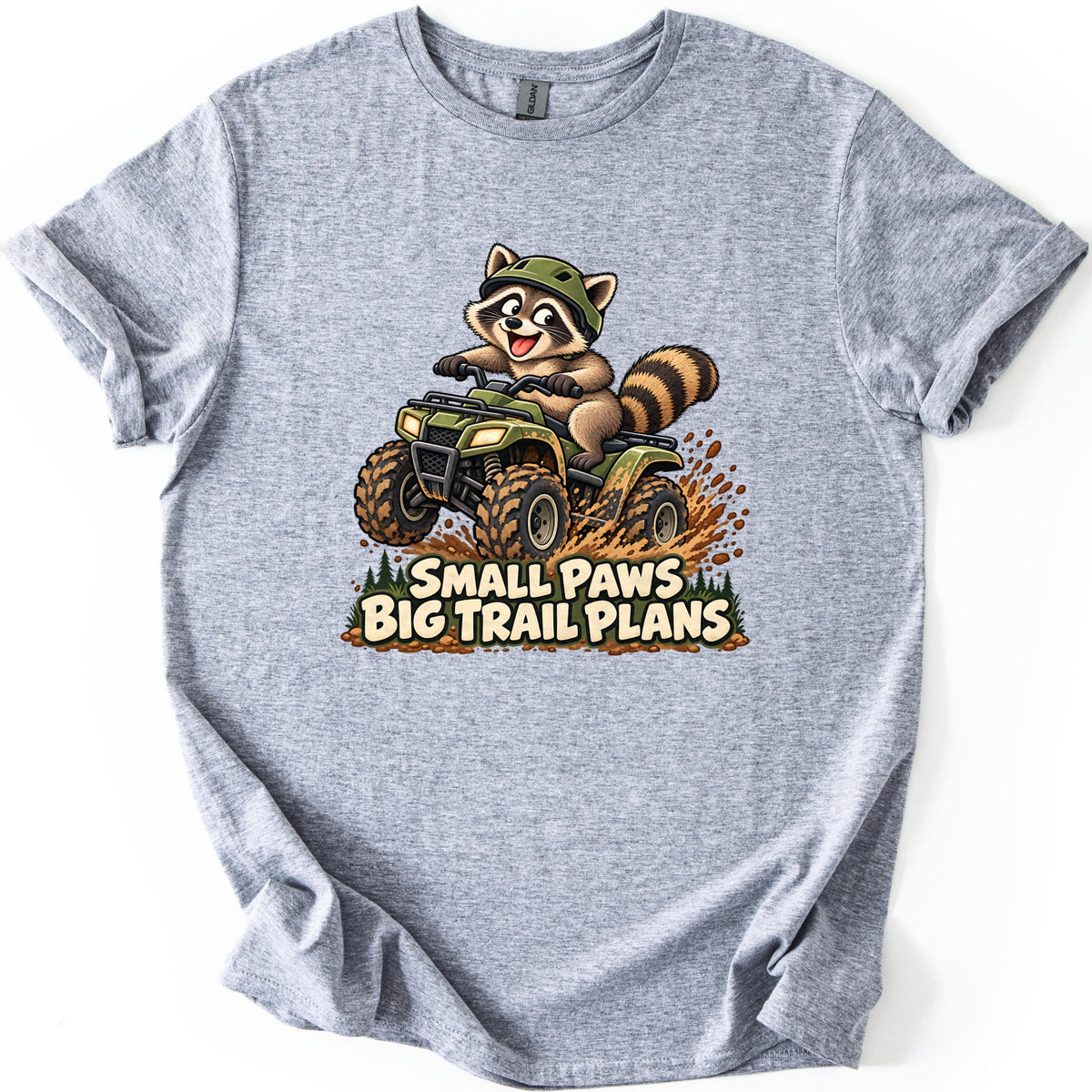 Small Paws Big Trail Plans Comfort Colors T-shirt in Sport Grey, laid flat. A cartoon raccoon in a green helmet rides a muddy green ATV, kicking up dirt beside pine trees, above the words Small Paws Big Trail Plans. The shirt lies on a white background with cuffed sleeves.