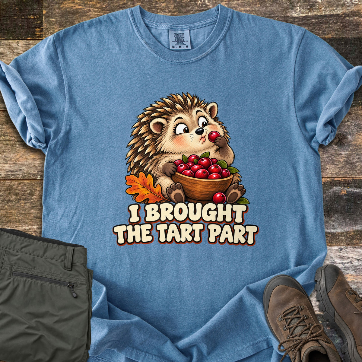 I Brought the Tart Part Comfort Colors T-shirt in Blue Jean, laid flat. A cartoon hedgehog nibbles a cranberry while holding a wooden bowl of cranberries beside an orange oak leaf, above the words I Brought the Tart Part. Olive pants and brown hiking boots sit on a wood background.