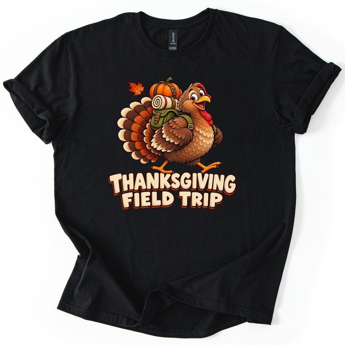 Thanksgiving Field Trip Comfort Colors T-shirt in Black, laid flat. A smiling cartoon turkey walks with a green backpack topped by a rolled blanket and a pumpkin, beside a falling maple leaf, above the words Thanksgiving Field Trip. The sleeves are cuffed on a white surface.