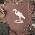 Great Blue Heron Comfort Colors T-shirt in Espresso, laid flat. A cream silhouette of a heron wading with its beak open, one foot raised, beside cattails over a patch of water. Sleeves rolled, on weathered wood with olive hiking pants and brown hiking boots.