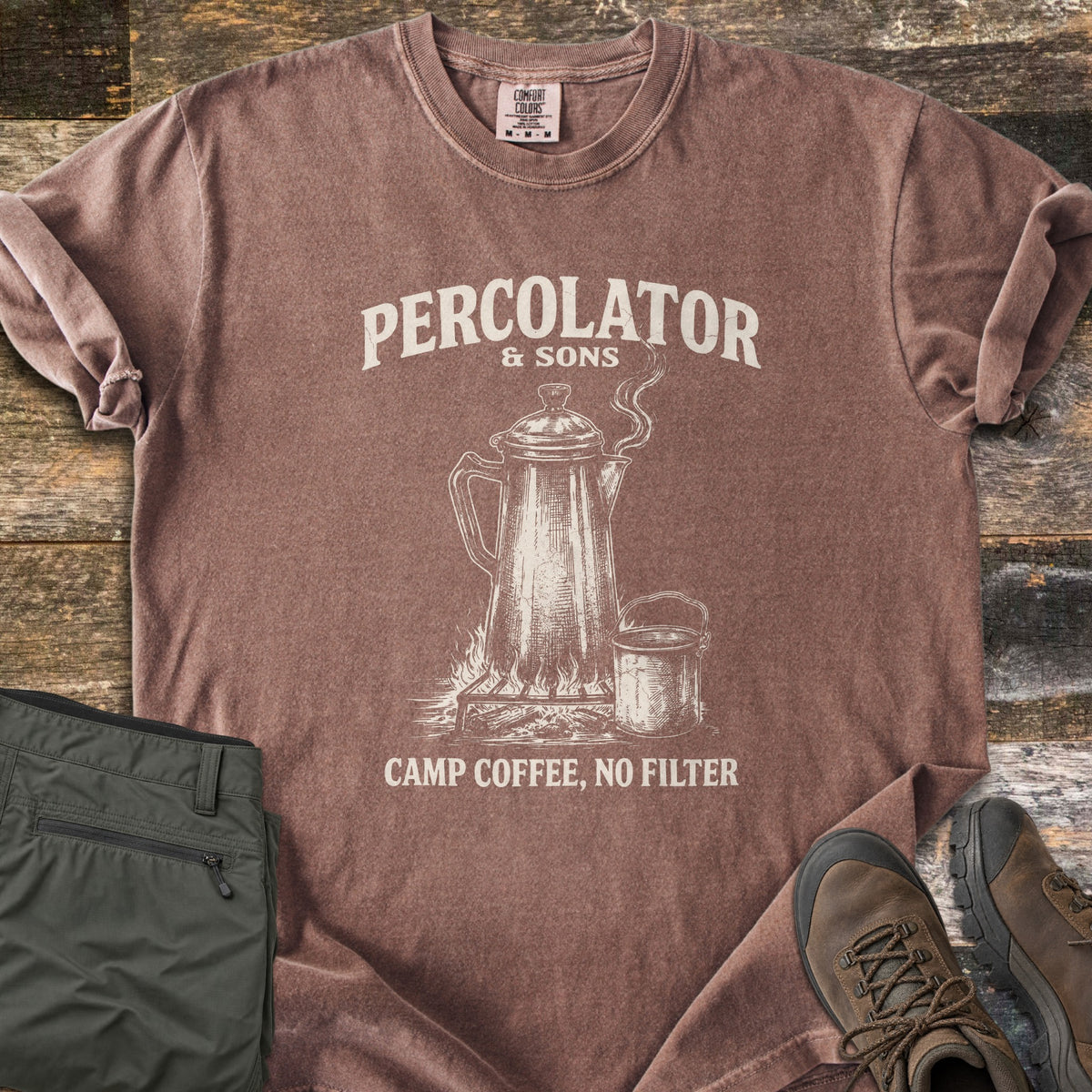 Percolator & Sons Comfort Colors T-shirt in Espresso, laid flat. Cream ink drawing of a steaming percolator and tin mug on a campfire grate, under the words Percolator & Sons and above Camp Coffee, No Filter. Olive pants and brown hiking boots lie on weathered wood.