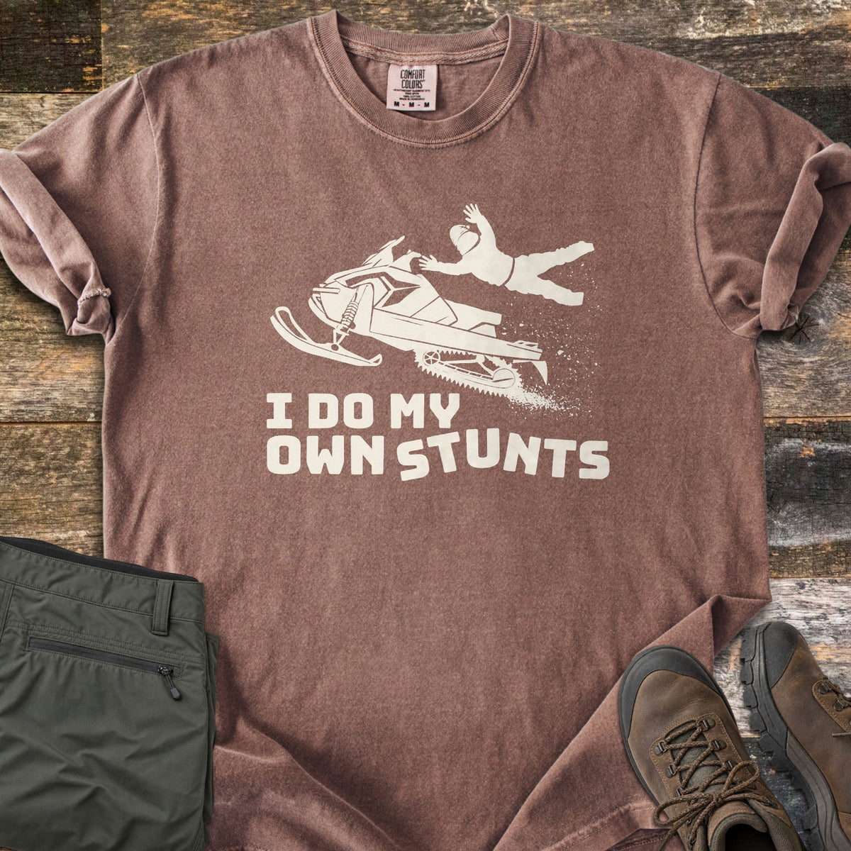 I Do My Own Stunts Comfort Colors T-shirt in Espresso, laid flat. A cream silhouette of a rider flying off a jumping snowmobile, snow spraying behind, above the words I Do My Own Stunts. Folded olive cargo pants and brown hiking boots sit beside it on weathered wood.