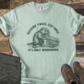 Measure Twice Beaver Comfort Colors T-shirt in Bay, laid flat. A green sketch of a beaver gnawing a pile of logs, with Measure Twice, Cut Once arched above and It's Only Woodwork below. Folded olive cargo pants and brown hiking boots sit on a weathered wood surface.