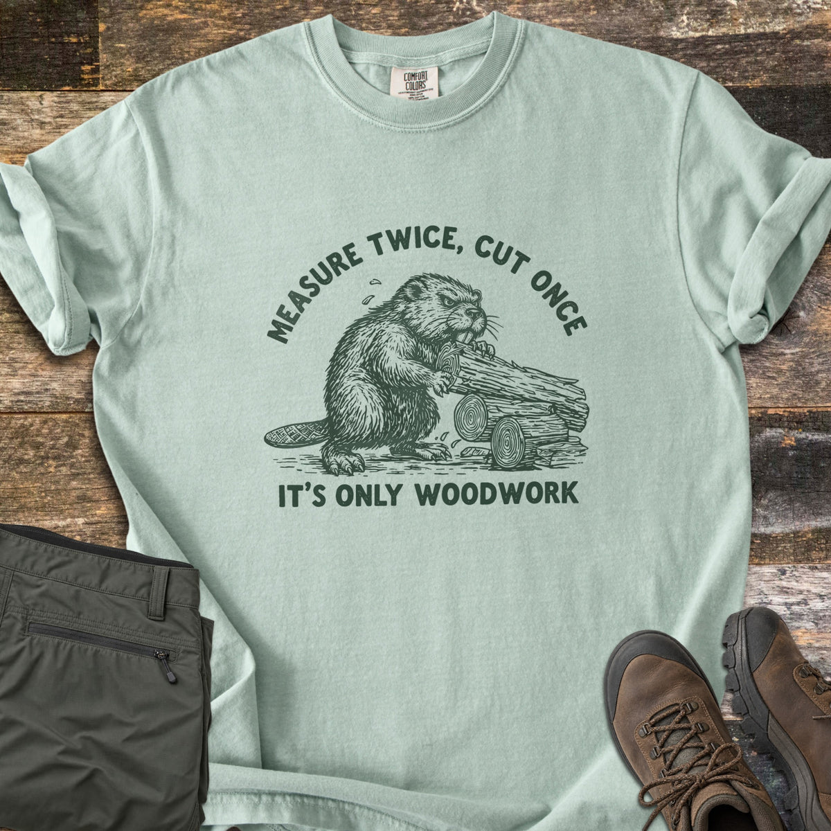 Measure Twice Beaver Comfort Colors T-shirt in Bay, laid flat. A green sketch of a beaver gnawing a pile of logs, with Measure Twice, Cut Once arched above and It's Only Woodwork below. Folded olive cargo pants and brown hiking boots sit on a weathered wood surface.