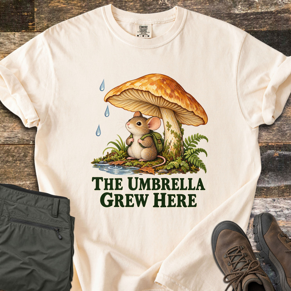The Umbrella Grew Here Comfort Colors T-shirt in Ivory, laid flat. A mouse wearing a green backpack shelters under a large orange mushroom from falling raindrops, above the words The Umbrella Grew Here. Olive pants and brown hiking boots lie beside it on weathered wood.