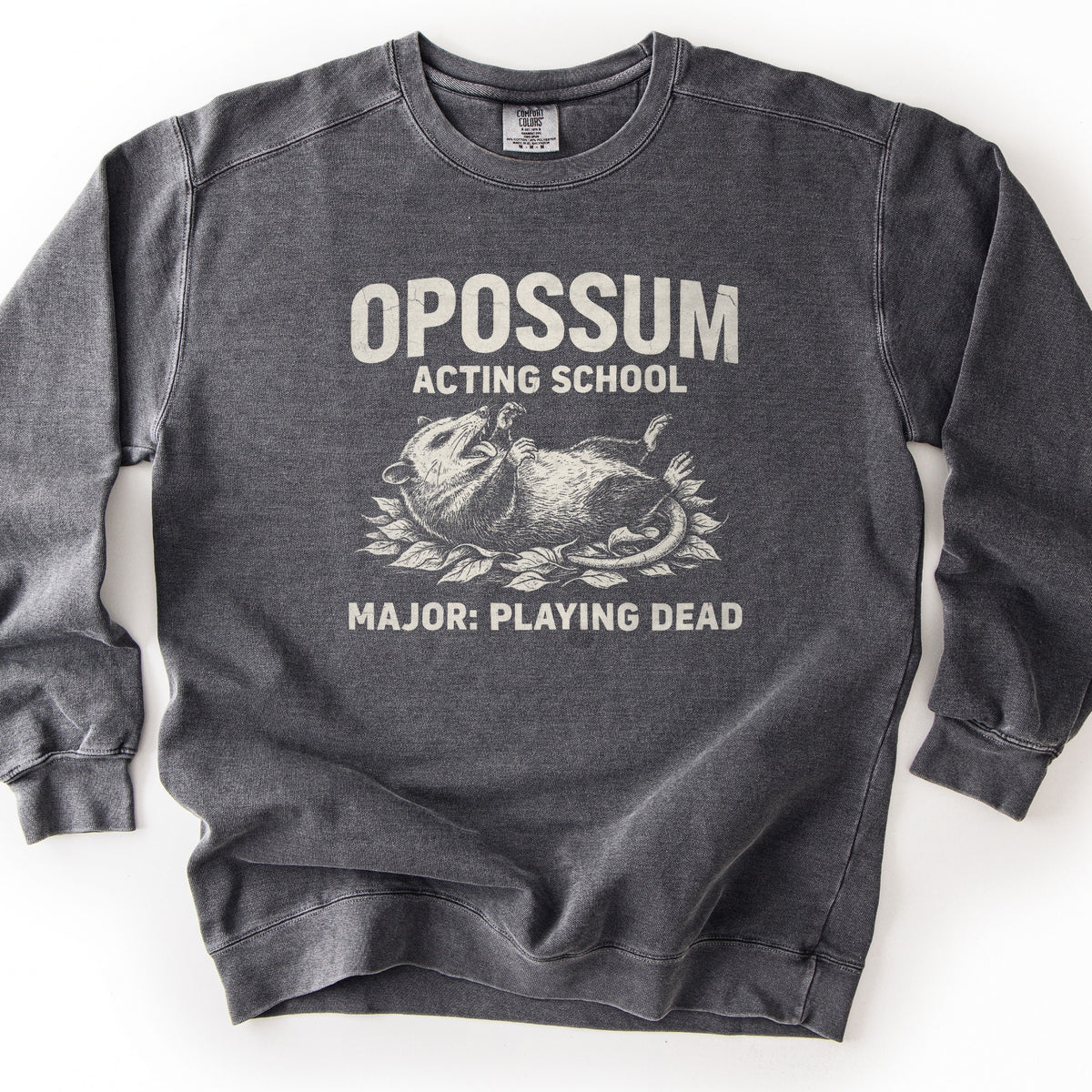 Opossum Acting School Comfort Colors crewneck sweatshirt in Pepper, laid flat. Cream print of a sketched opossum lying on its back in leaves, mouth open, under the words Opossum Acting School and above Major: Playing Dead. The dark gray sweatshirt lies on a white surface.