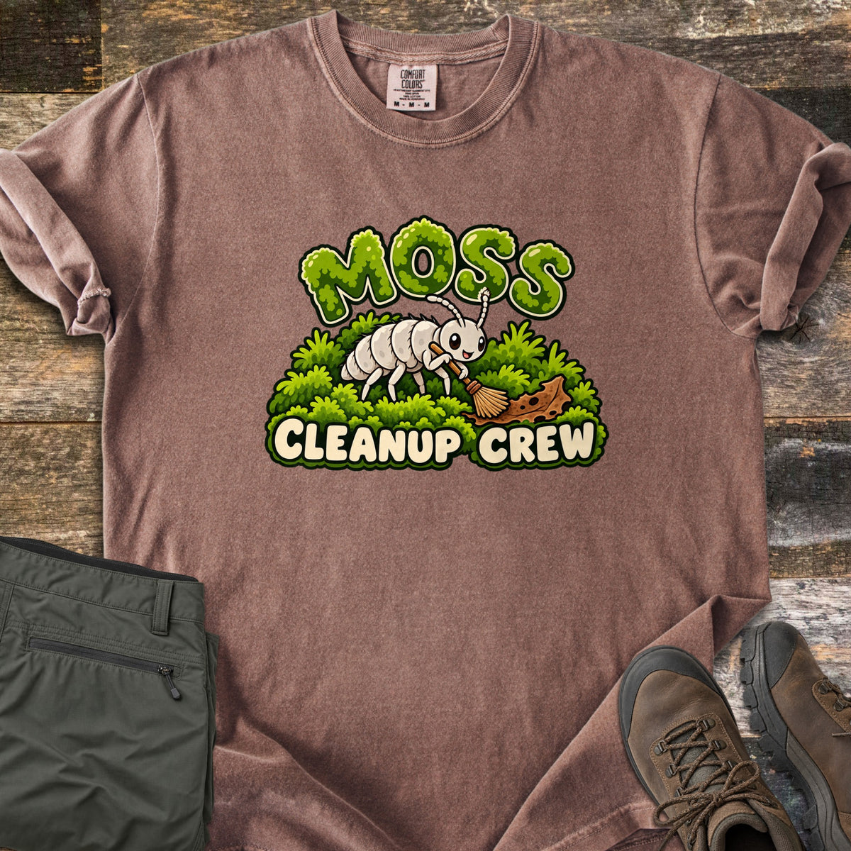 Moss Cleanup Crew Comfort Colors T-shirt in Espresso, laid flat. A smiling white cartoon bug sweeps a brown leaf with a broom atop green moss, below the word MOSS in mossy letters and above CLEANUP CREW. It lies on weathered wood with folded olive pants and brown hiking boots.
