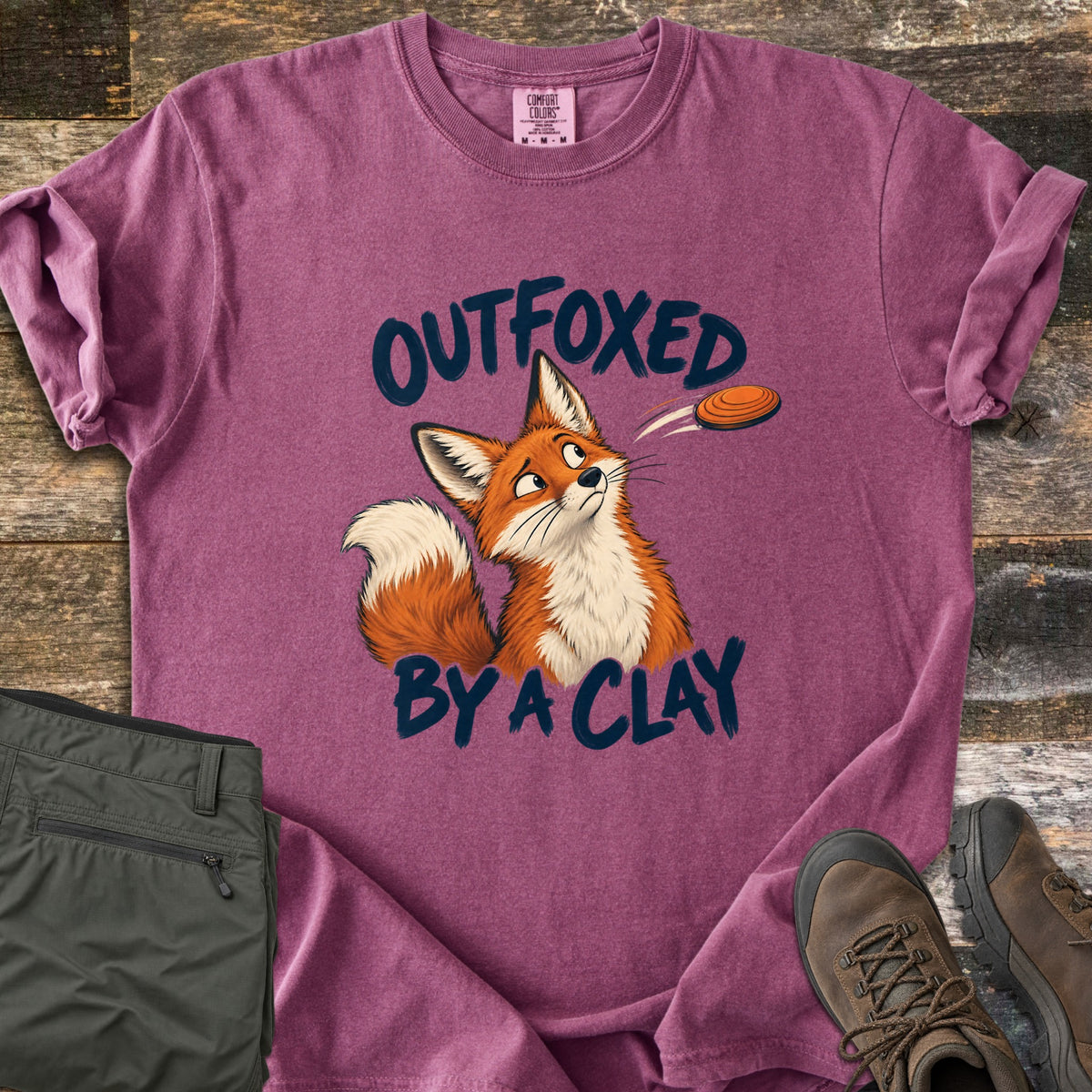 Outfoxed by a Clay Comfort Colors T-shirt in Berry, laid flat. A cartoon red fox glances sideways at an orange clay target flying past, between the words Outfoxed By A Clay in navy brush letters. Sleeves rolled on rustic wood with olive pants and brown hiking boots.