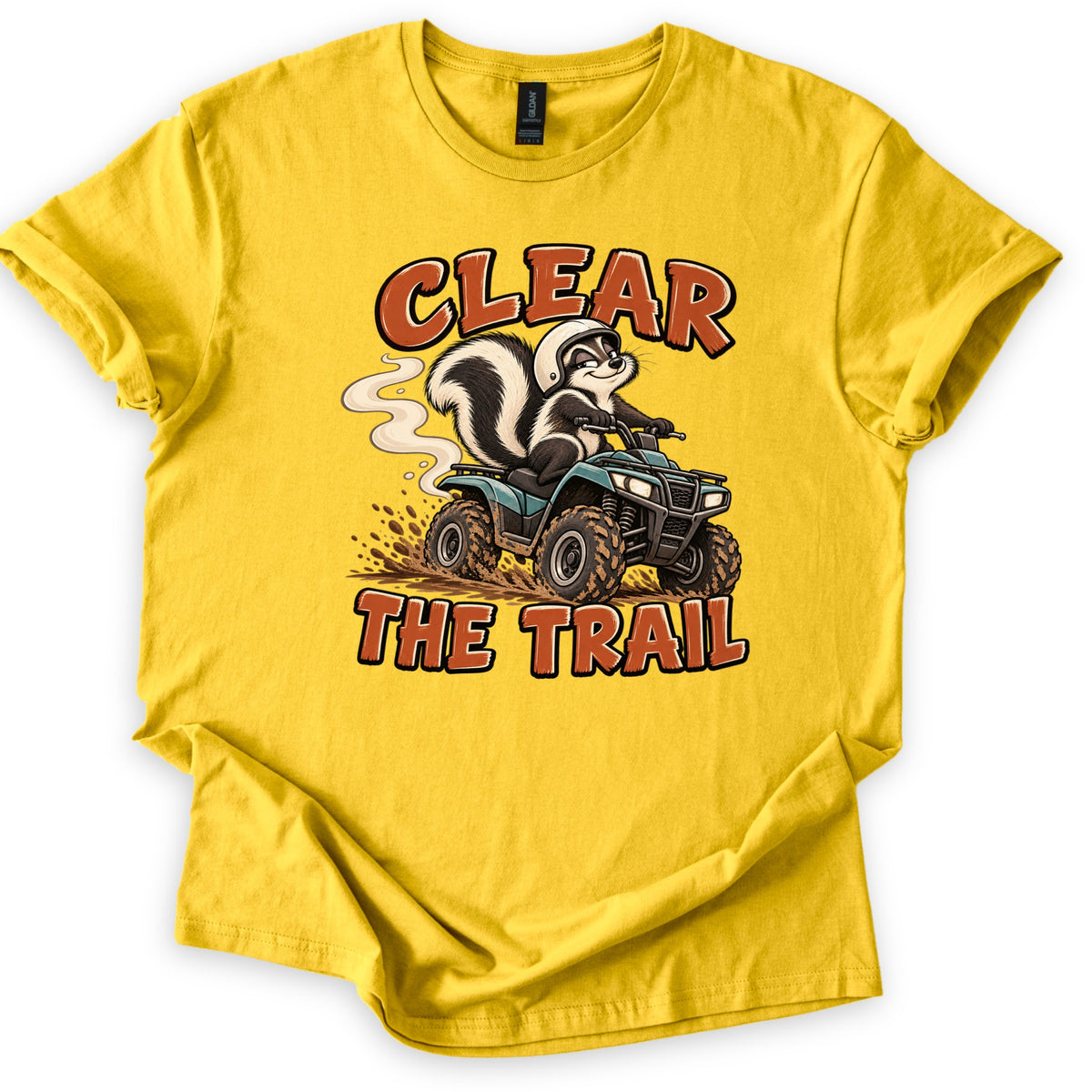 Clear the Trail Comfort Colors T-shirt in Daisy, laid flat. A cartoon skunk in a white helmet drives a teal ATV through splashing mud with a smoke trail behind it, framed by the words Clear The Trail in rust-orange letters. The shirt lies on a white background.