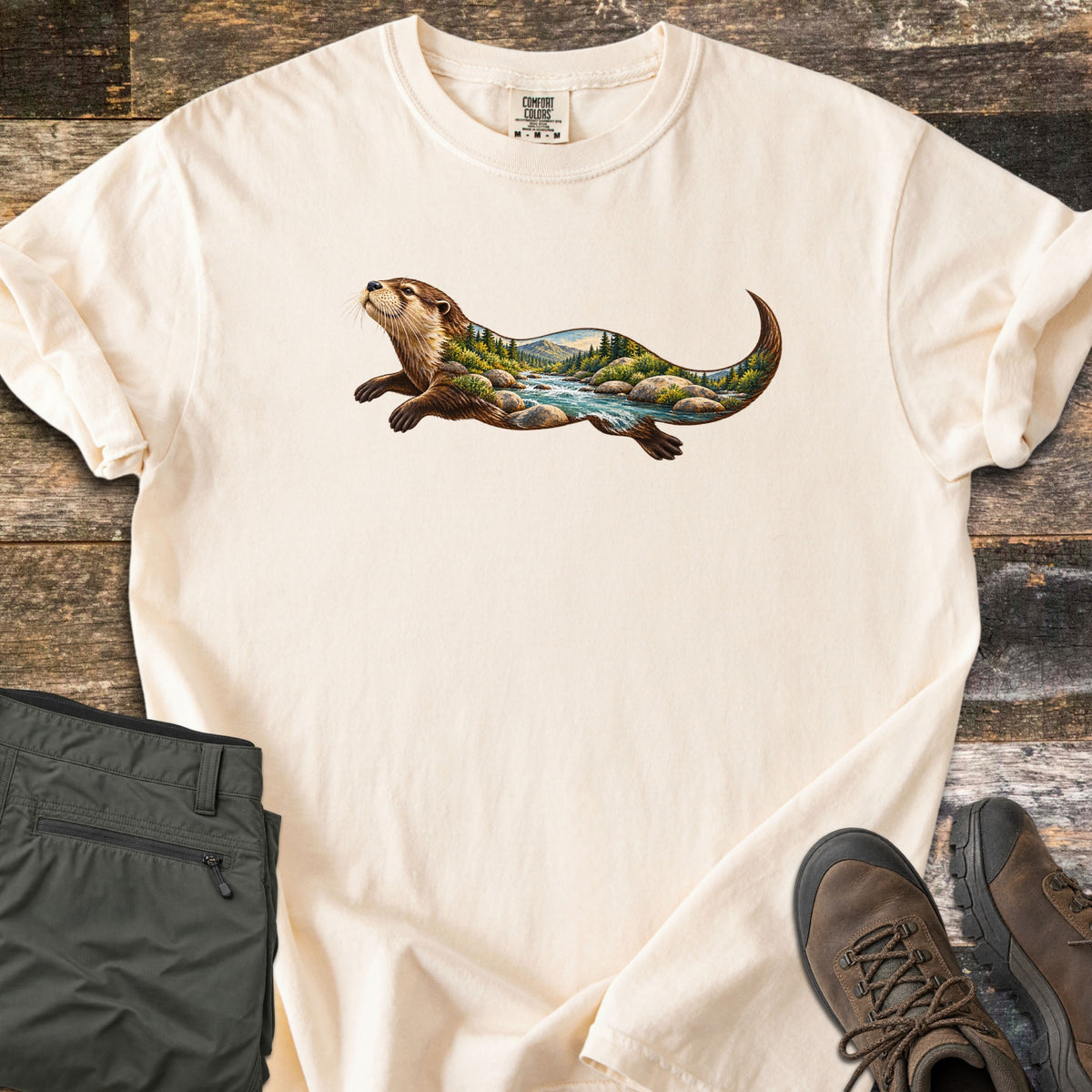 Otter River Bend Comfort Colors T-shirt in Ivory, laid flat. A river otter swims across the chest, its body filled with a mountain river scene of rushing water, boulders and pine trees. Olive pants and brown hiking boots lie beside it on weathered wood planks.
