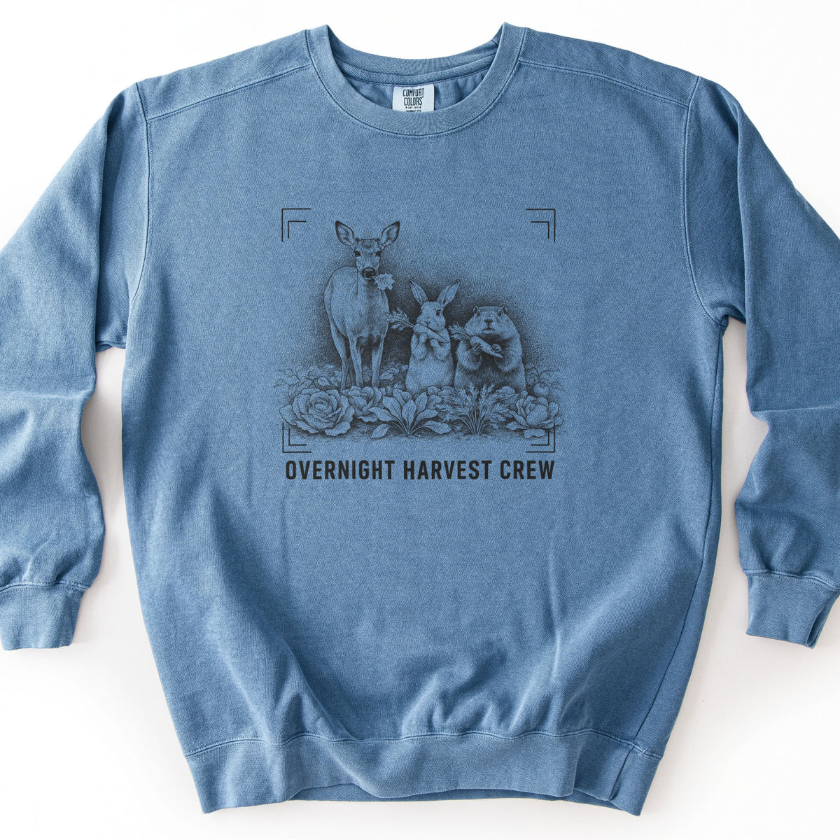 Overnight Harvest Crew Comfort Colors crewneck sweatshirt in Blue Jean, laid flat. A dark ink sketch of a deer with a leaf in its mouth beside a rabbit and a groundhog holding carrots among cabbages, within corner brackets above the words OVERNIGHT HARVEST CREW. On a white surface.