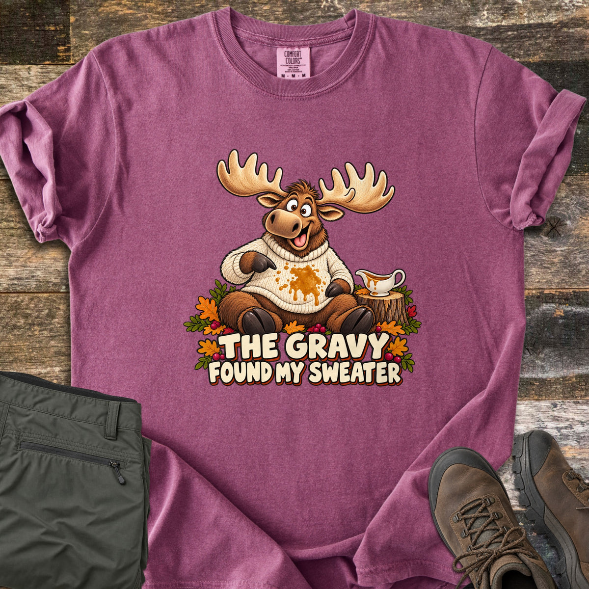 The Gravy Found My Sweater Comfort Colors T-shirt in Berry, laid flat. A cartoon moose in a cream knit sweater points at a gravy stain beside a gravy boat on a stump, above the words The Gravy Found My Sweater. Olive pants and brown boots sit on a wood floor.