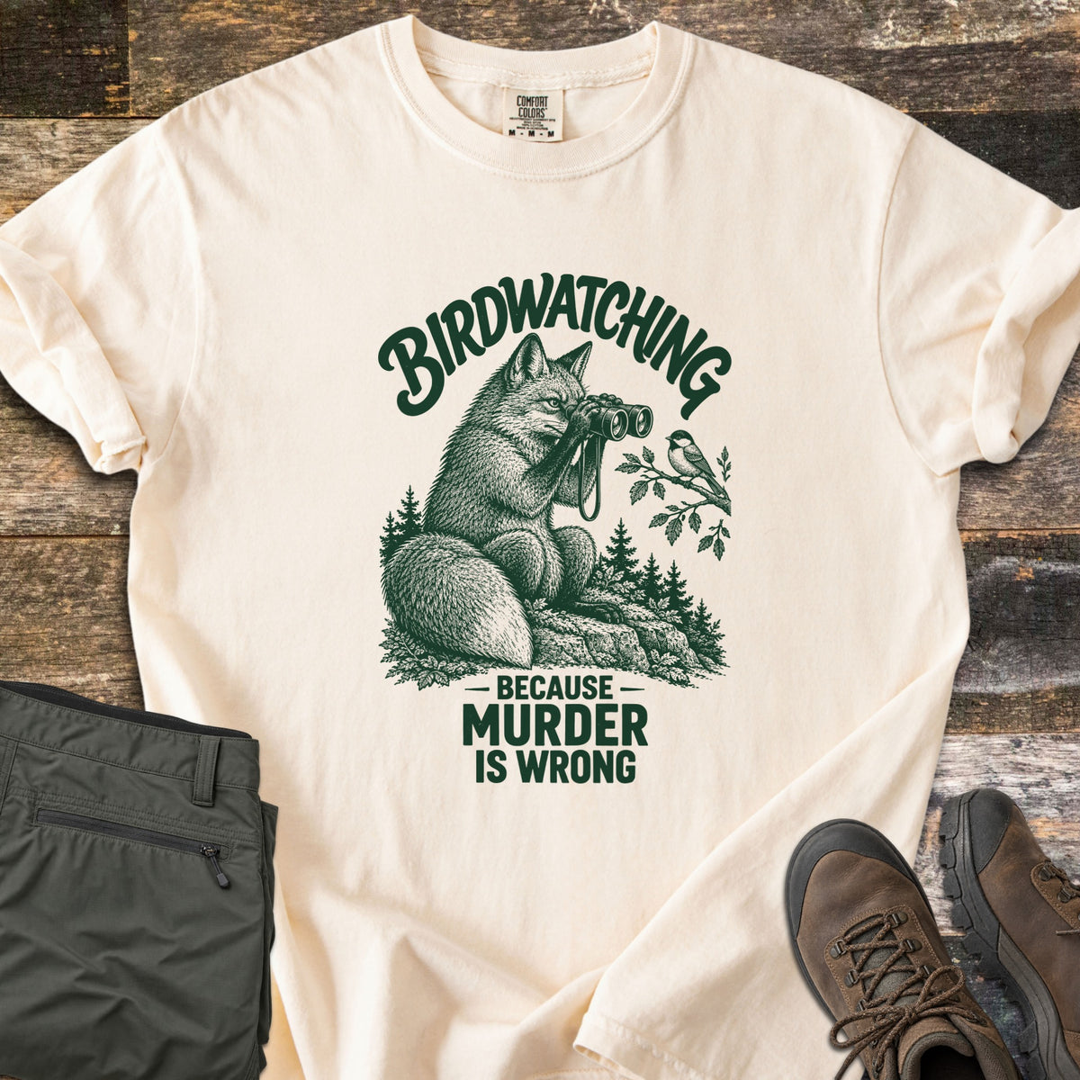 Birdwatching Because Murder Is Wrong Comfort Colors T-shirt in Ivory, laid flat. A fox on a rock looks through binoculars at a small bird on a branch, with the arched word Birdwatching above and Because Murder Is Wrong below, in dark green ink. Olive hiking pants and brown boots lie on weathered wood.