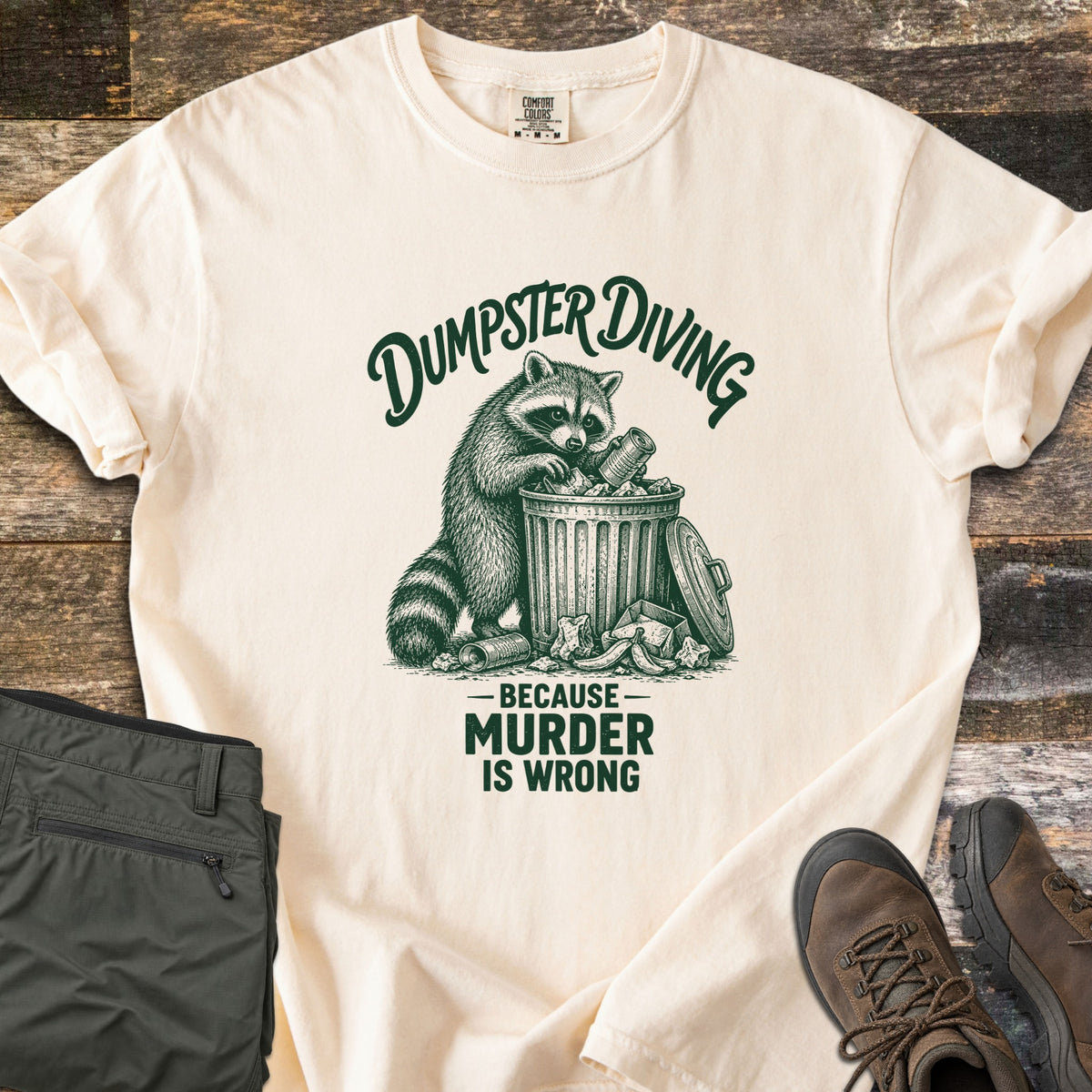 Dumpster Diving Because Murder Is Wrong Comfort Colors T-shirt in Ivory, laid flat. A dark green sketch of a raccoon digging through an overflowing metal trash can, with the words Dumpster Diving above and Because Murder Is Wrong below. Olive cargo pants and brown hiking boots lie on a wood floor.