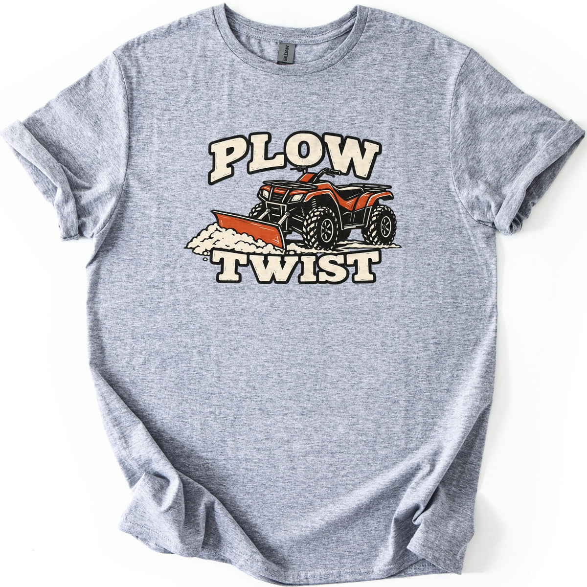 Plow Twist Comfort Colors T-shirt in Sport Grey, laid flat. An orange ATV with a front plow blade pushes a pile of snow, with the words Plow Twist in cream block letters above and below. The shirt lies on a white background with the sleeves cuffed.