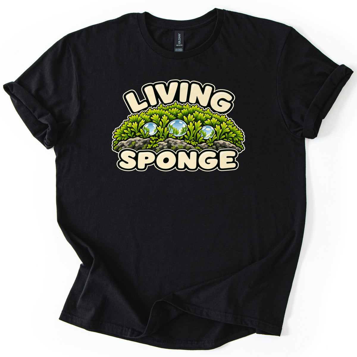 Living Sponge Comfort Colors T-shirt in Black, laid flat. Cream bubble letters spell LIVING above and SPONGE below a cartoon mound of green moss on gray rocks with three shiny water droplets. The shirt lies on a white surface with sleeves rolled.