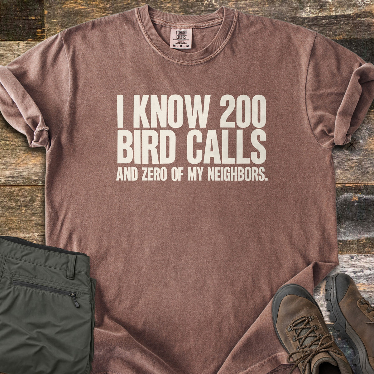 I Know 200 Bird Calls and Zero of My Neighbors Comfort Colors T-shirt in Espresso, laid flat. Bold off-white capital letters read I KNOW 200 BIRD CALLS AND ZERO OF MY NEIGHBORS. The shirt has rolled sleeves and lies on weathered wood boards beside folded olive hiking pants and brown hiking boots.