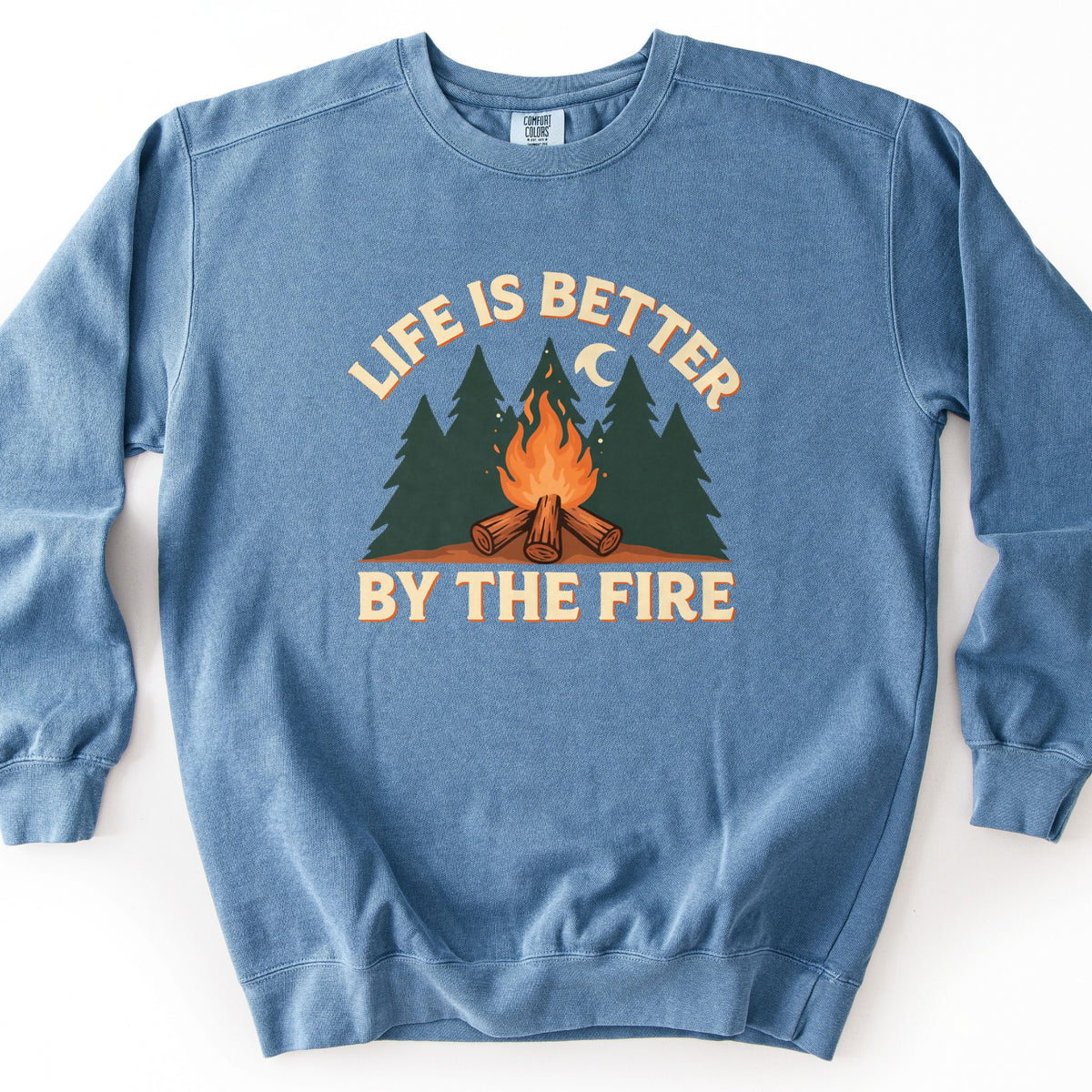 Life Is Better By The Fire Comfort Colors crewneck sweatshirt in Blue Jean, laid flat. Cream letters with an orange shadow arch Life Is Better over dark green pine trees, a crescent moon and a campfire of crossed logs, with By The Fire below. It lies on a white surface.